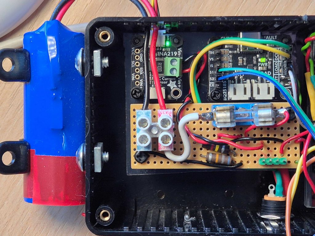 Photograph shows how the Adafruit charger and current detection modules have been mounted inside a standard plastic enclosure along with a small piece of stripboard that has a choc-block connector (2 channel), fuse and a potential divider consisting of 2 resistors in series which can be connected to an analogue pin on the Arduino as an alternative method for monitoring battery voltage. The 26650 LFP battery can be seen held in place by two P clips on the outside of the plastic enclosure.